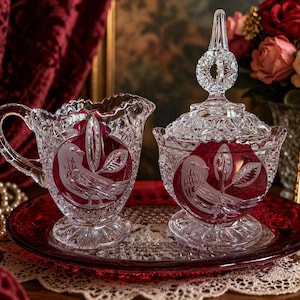 May include: A crystal creamer and covered sugar bowl set with a red interior and bird design. The set sits on a red glass tray. The creamer has a handle, and the sugar bowl has a decorative lid.