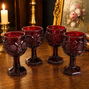 May include: Four ruby red wine glasses with embossed patterns. The glasses have a short stem and a hexagonal base. A lit candle and antique decor are in the background.