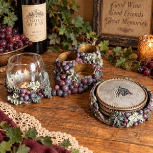 May include: A set of decorative home accessories featuring a glass candle holder, a triple-votive candle holder, and a stack of coasters, all adorned with sculpted purple grapes and green leaves. The coasters feature a central grape cluster design.