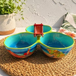 Peut inclure: Un bol de service en céramique à trois compartiments avec un intérieur turquoise vif et un extérieur peint à la main représentant une scène de plage avec une chaise longue rouge, un cocktail sur une table et un parasol à fleurs jaunes.