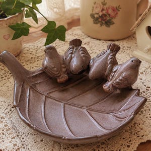 May include: A brown ceramic dish shaped like a large leaf with four small bird figurines perched along the curved edge. The dish features a textured surface with visible veins and a glossy finish, providing a decorative tray for small items.