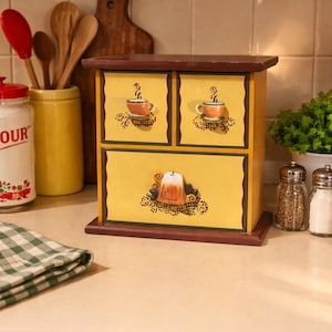 Peut inclure: Un coffret de rangement en bois avec un dessus et une base marron foncé, doté de trois tiroirs jaunes. Les deux petits tiroirs supérieurs ont des boutons en céramique en forme de tasse, et le grand tiroir inférieur a un bouton en théière.
