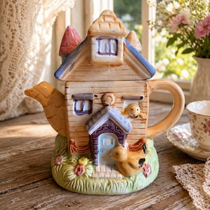 Op de afbeelding: Een grillige keramische theepot in de vorm van een huis, met een lichtbruine buitenkant en blauwe en roze accenten. De theepot heeft een eendenfiguur en een hond die uit een raam kijkt. De basis is groen met bloemendetails. Een charmante toevoeging aan elke theeset.
