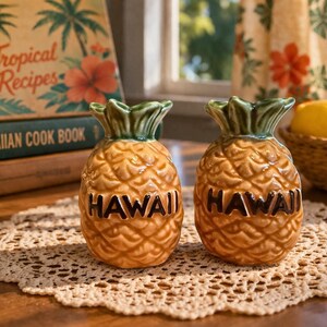 May include: Two ceramic pineapple-shaped salt and pepper shakers with the word "HAWAII" in black lettering. The shakers are on a cream-coloured lace doily, with a stack of cookbooks in the background.