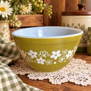 May include: A round mixing bowl with a smooth olive green exterior featuring a repeating white floral pattern of five-petaled flowers and small dots. The interior of the bowl is a solid, light blue colour with a wide, circular rim.