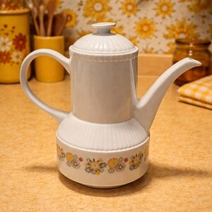 May include: A white ceramic mug with a black handle and the text "But First, Coffee" in a bold, black font. The mug is set against a blurred background. The mug appears to be approximately 10 cm tall.