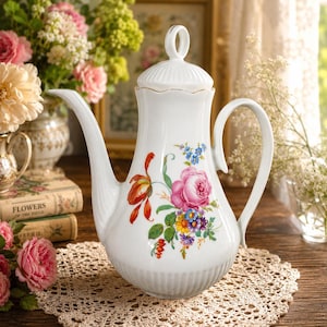 Puede incluir: Una cafetera de porcelana blanca con base acanalada y tapa con remate en forma de anillo. El cuerpo está decorado con un colorido ramo de flores que incluye una gran rosa rosa, un tulipán naranja y pequeñas flores azules y amarillas.