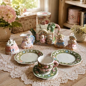 May include: A ceramic tea set featuring a cottage-shaped teapot, a creamer, two small plates, a teacup with a saucer, and two bear figurines. The pieces are decorated with pastel colours, floral patterns, and sculpted bows, creating a cohesive set.
