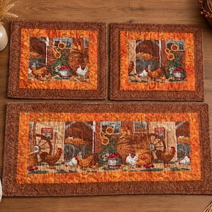May include: Two rectangular quilted placemats and one long quilted table runner featuring a farm scene with chickens, sunflowers, and wooden wagon wheels. The fabric has a warm colour palette of orange, brown, and gold with a textured border.