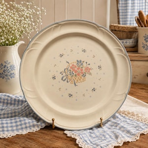 Peut inclure: Une assiette ronde en céramique avec une base couleur crème et un bord bleu clair. Le centre présente une illustration décorative d'un panier tressé rempli de fleurs roses et bleues, entouré de petits motifs floraux bleus dispersés.