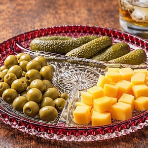 May include: A divided serving dish with olives, pickles, and cheese cubes. The red-rimmed glass dish is filled with green olives, dill pickles, and yellow cheese. A glass of amber liquid is in the background.
