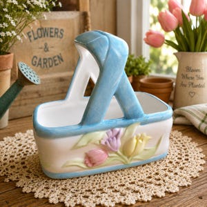 Peut inclure: Un panier en céramique blanche avec une anse et un bord bleus, orné d'un motif en relief de tulipes roses, violettes et jaunes sur le devant. Le panier possède une ouverture supérieure et une base rectangulaire, avec une finition lisse et brillante.