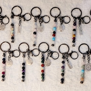 May include: A collection of twelve metal keychains, each featuring a circular ring, a lobster clasp, a vertical strand of multi-colored beads, and a dangling metal charm engraved with various inspirational phrases and symbols.