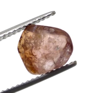 May include: A translucent, light brown gemstone with a rounded shape, held by a pair of silver tweezers. The gem has a slightly irregular surface and a subtle, warm color gradient, ranging from pale pink to deeper brown.