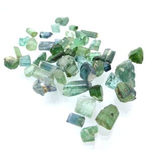 May include: A collection of green and blue gemstones, some with a slight shimmer, scattered on a white surface. The gemstones are various shapes and sizes, with some being long and thin, while others are more rounded.