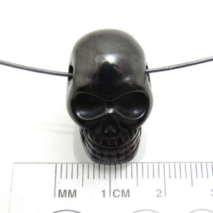 May include: Black skull bead pendant made of polished hematite. The bead is approximately 1 cm in diameter and has a hole drilled through the top for stringing.