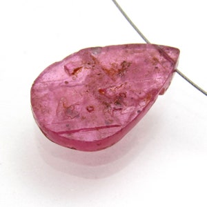 May include: A pink teardrop-shaped gemstone pendant with a wire loop for hanging. The gemstone has a rough, textured surface and appears to be carved with a design.