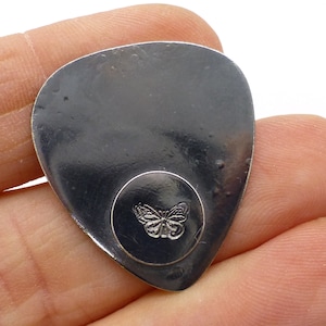 May include: A silver guitar pick with a circular design featuring a butterfly in the centre.