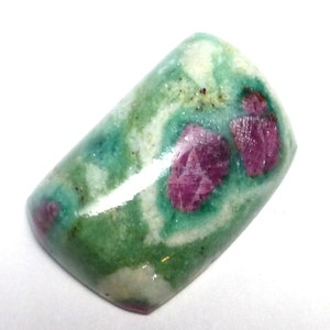 May include: A green and white cabochon gemstone with pink ruby inclusions. The gemstone is oval shaped and has a smooth, polished surface.
