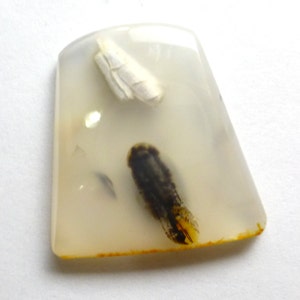 May include: A white agate cabochon with black and yellow inclusions. The cabochon is translucent and has a smooth, polished surface.