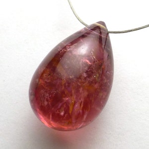 May include: A teardrop-shaped, translucent gemstone pendant. The pendant is a deep reddish-pink color with internal veining and inclusions. A thin, silver-colored wire is attached at the top for hanging. The gemstone has a polished, glossy finish.