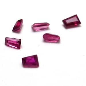 May include: A collection of six faceted, rectangular-shaped gemstones in varying sizes. The gems are a deep, vibrant magenta color, with a polished, reflective surface. The gemstones are scattered on a white background.