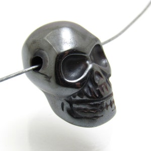 May include: A black hematite skull bead with a hole drilled through the top for stringing. The bead is carved with detailed facial features, including eye sockets, a nose, and a mouth.