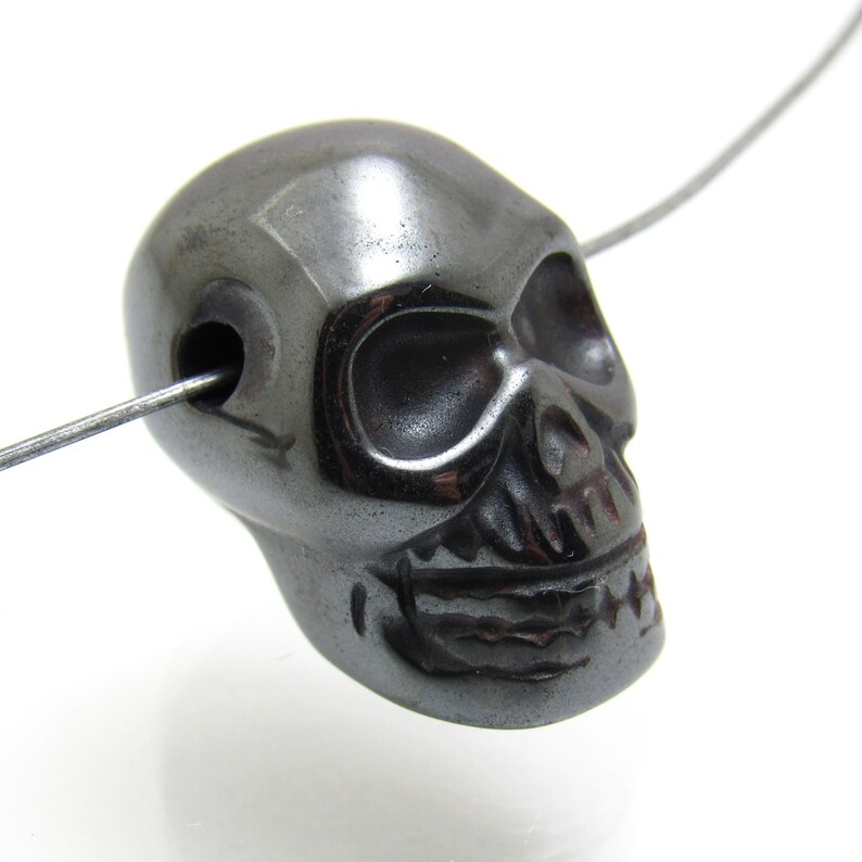 May include: A black hematite skull bead with a hole drilled through the top for stringing. The bead is carved with detailed facial features, including eye sockets, a nose, and a mouth.