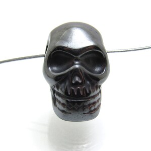 May include: A black skull bead made of hematite. The bead is detailed with a skull face and teeth.