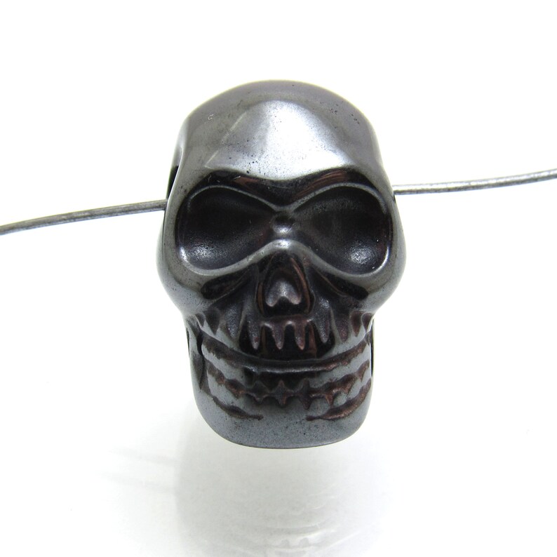 May include: A black skull bead made of hematite. The bead is detailed with a skull face and teeth.