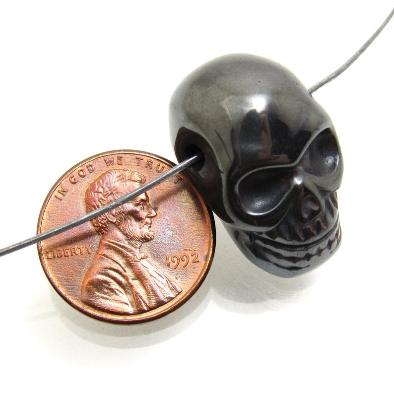 May include: A black skull bead made of hematite, next to a 1992 US penny for size comparison. The bead is smooth and polished, with a shiny finish.