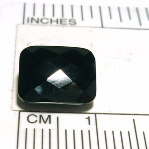 K&ouml;nnte beinhalten: Ein dunkler, facettierter, schwarzer Edelstein mit quadratischer Form. Der Edelstein ist etwa 1 cm breit und liegt auf einer wei&szlig;en Oberfl&auml;che mit einem Lineal.