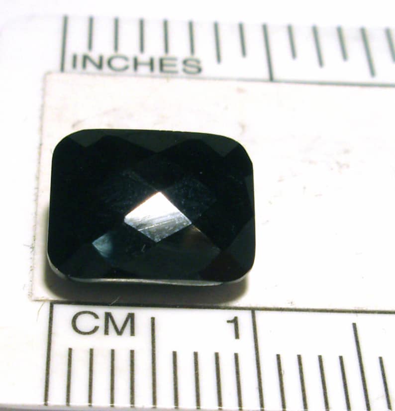K&ouml;nnte beinhalten: Ein dunkler, facettierter, schwarzer Edelstein mit quadratischer Form. Der Edelstein ist etwa 1 cm breit und liegt auf einer wei&szlig;en Oberfl&auml;che mit einem Lineal.