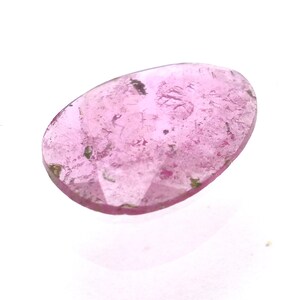 May include: A polished, oval-shaped pink gemstone with a translucent appearance. The gem has a light pink hue with darker pink and green inclusions. The stone is set against a white background, highlighting its color and clarity.