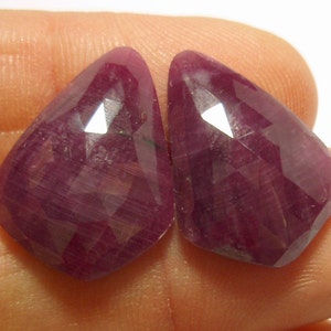 May include: Two faceted, triangular-shaped, deep red gemstones. The gems have a polished, reflective surface with multiple facets that catch the light. The color is a rich, dark red, with a slight variation in tone across the surface. These gems are likely rubies.
