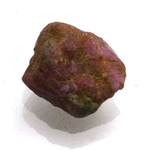 May include: A rough, dark pink ruby gemstone with a slightly irregular shape. The ruby has a dull, matte finish and is covered in small, brown inclusions.