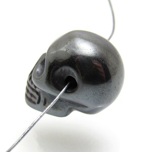 May include: A black skull bead made of hematite, a type of iron ore, with a hole drilled through the top for stringing.
