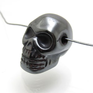 May include: A black hematite skull bead with a detailed carving of a skull face. The bead has a hole drilled through the top for stringing.