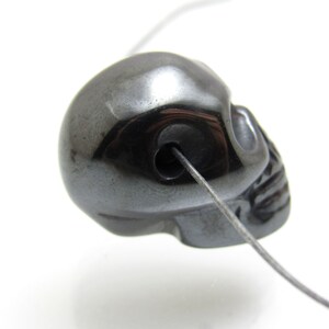 May include: A black hematite skull bead with a hole drilled through the top for stringing. The bead is smooth and polished.