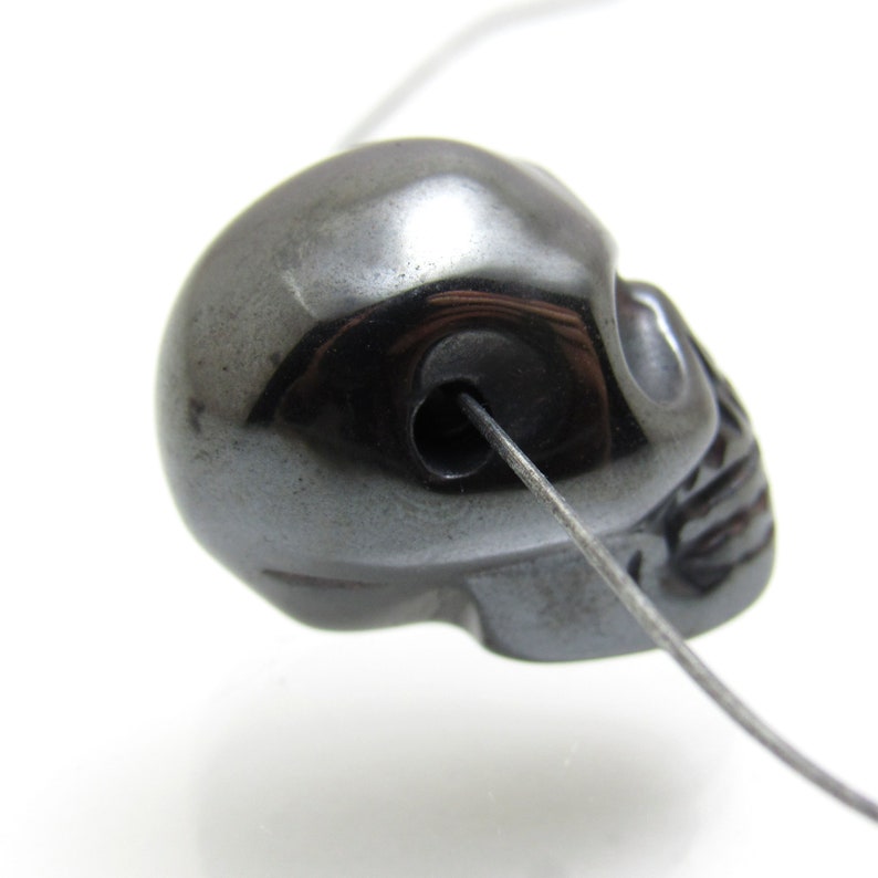 May include: A black hematite skull bead with a hole drilled through the top for stringing. The bead is smooth and polished.