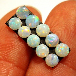 May include: A close-up of ten round, white opal gemstones with iridescent rainbow colors. The opals are arranged in a row on a black backing.
