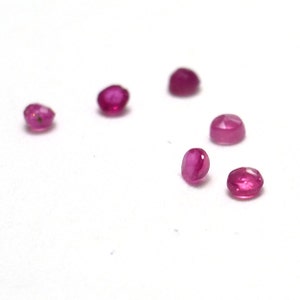 May include: Six small, round, pink ruby gemstones on a white surface.