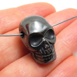 May include: A black hematite skull bead with a detailed carving of a skull face. The bead has a hole drilled through the top for stringing.