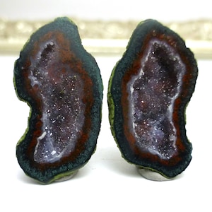 May include: Two geode halves with a purple and white crystal interior. The exterior is a dark green, red, and black color.