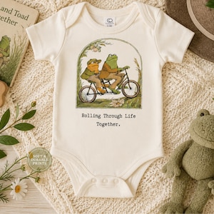May include: An off-white short-sleeve baby bodysuit featuring a graphic illustration of two frogs riding a tandem bicycle. Below the illustration, the text reads, Rolling Through Life Together. The garment has a round neckline and snap closures.