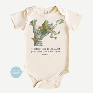 May include: A cream-colored short-sleeve baby bodysuit featuring a printed illustration of two frogs sitting in a tree reading books. Below the illustration, the text reads: Together, even the smallest pond feels like a whole new world.