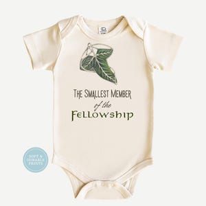 May include: A cream-colored short-sleeve baby bodysuit featuring a green leaf graphic and the text "THE SMALLEST MEMBER of the FELLOWSHIP" printed in black and green. A small circular blue badge in the corner reads "SOFT & DURABLE PRINTS".
