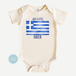 May include: Cream-colored baby bodysuit with the Greek flag design in blue and white. The text "JUST A LITTLE GREEK" is printed above and below the flag. The bodysuit has short sleeves and a round neckline.