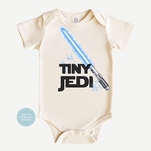 May include: Cream-colored baby onesie with the words "TINY JEDI" in black. A blue lightsaber graphic is featured on the front. The onesie has short sleeves and a round neckline. A small, round sticker with the text "SOFT & DURABLE PRINTS" is on the lower left.