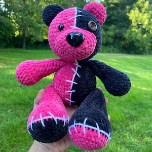 Puede incluir: Un oso de peluche hecho de hilo suave, con un diseño dividido en un lado de color rosa brillante y el otro en negro. Tiene una nariz de plástico negro brillante, un ojo de botón negro y detalles cosidos en blanco a lo largo de la costura.
